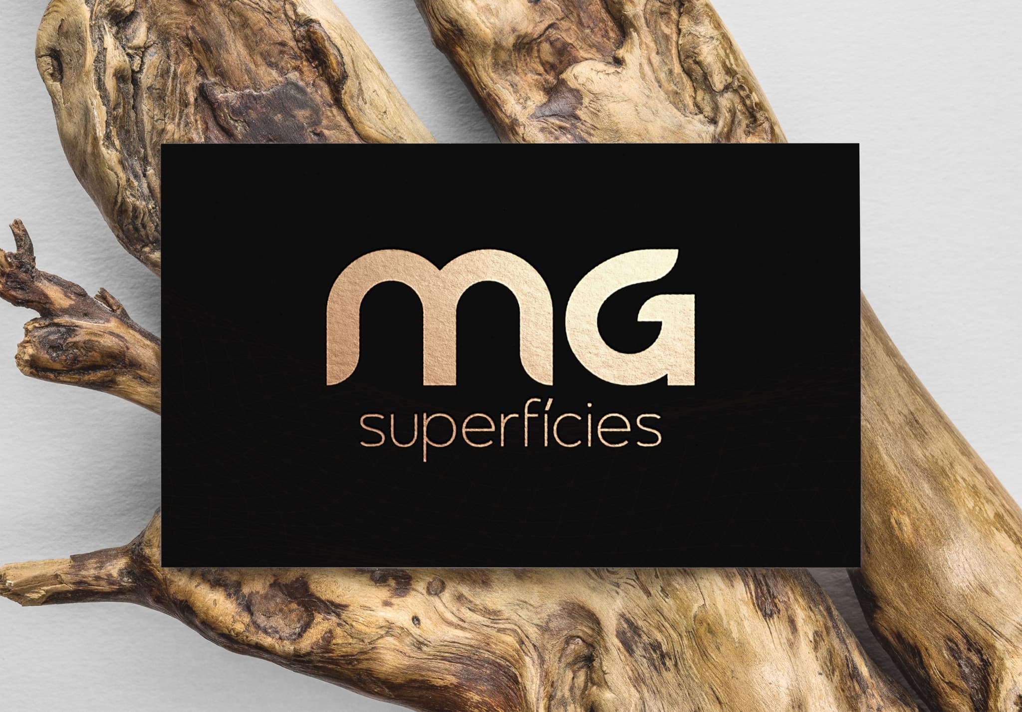 Cartão preto com a marca MG Superfícies em dourado sobre tronco de madeira