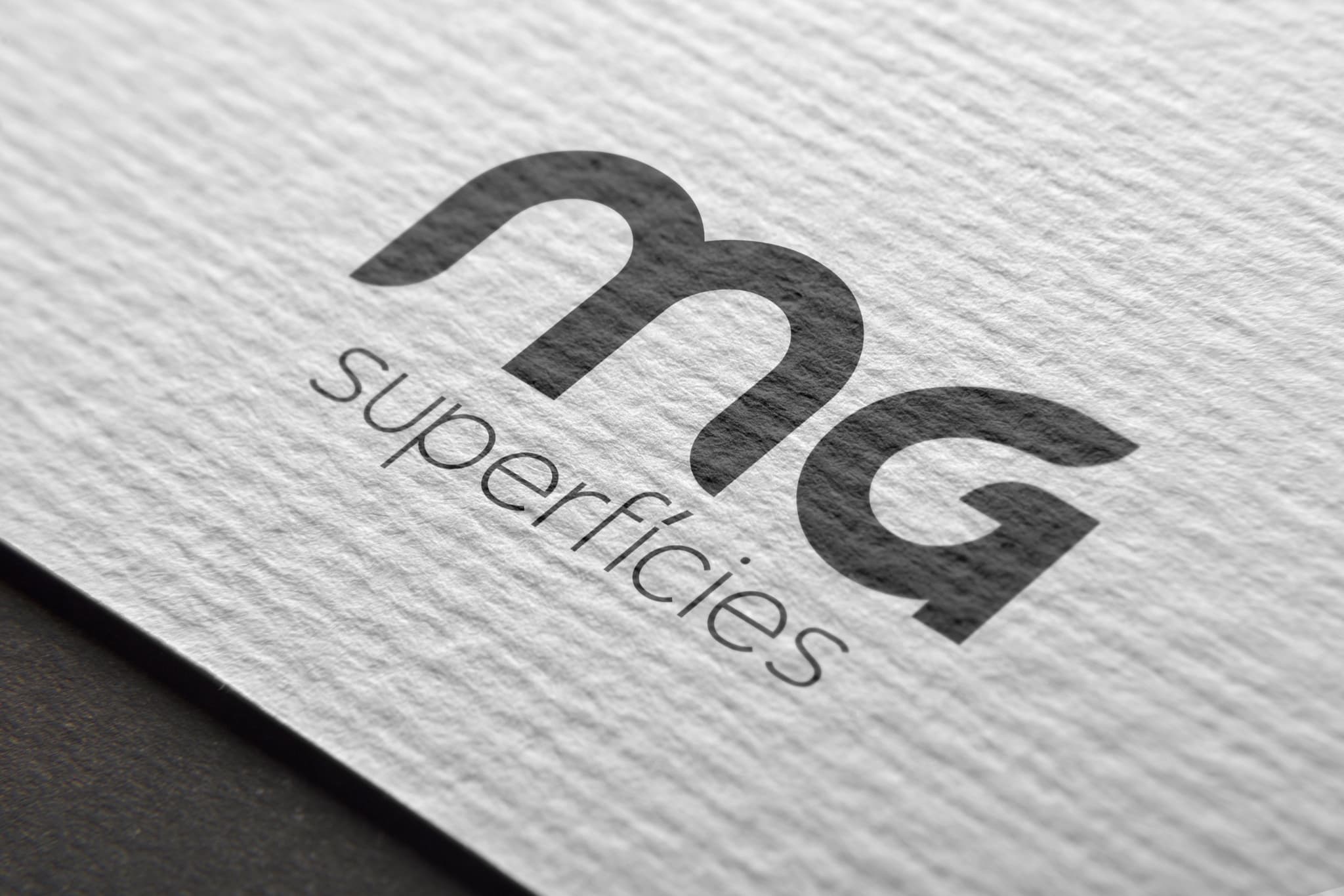 Logotipo MG Superfícies em relevo sobre papel texturizado cinza