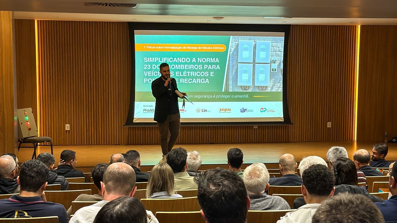 Palestrante no palco do 1º Fórum sobre Normatização de Recarga de Veículos Elétricos, visto do fundo do auditório lotado