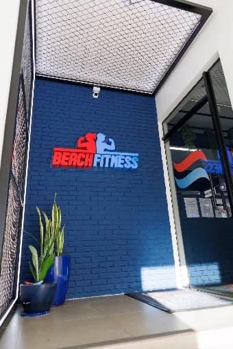 Entrada da Beach Fitness com o logotipo em relevo sobre parede de tijolo azul, ao lado de um vaso de espada-de-são-jorge