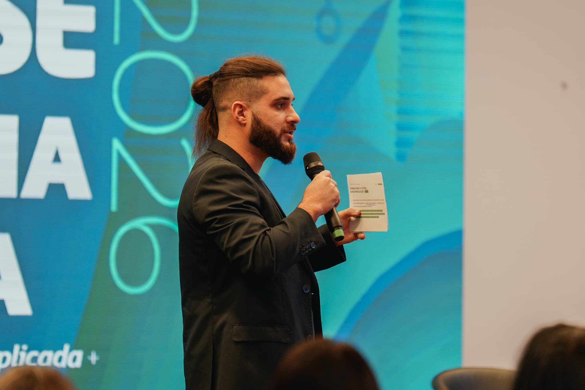 Mestre de cerimônia do CONTASE no palco com o microfone, diante do púlpito do evento e da mesa de convidados