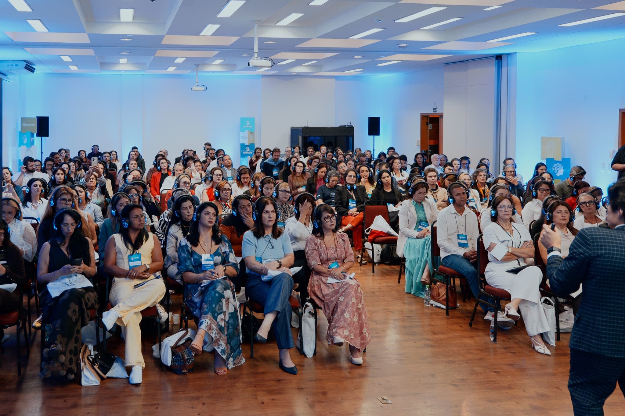 Auditório lotado do CONTASE visto do palco, com a plateia usando fones de tradução simultânea azuis e crachás do evento