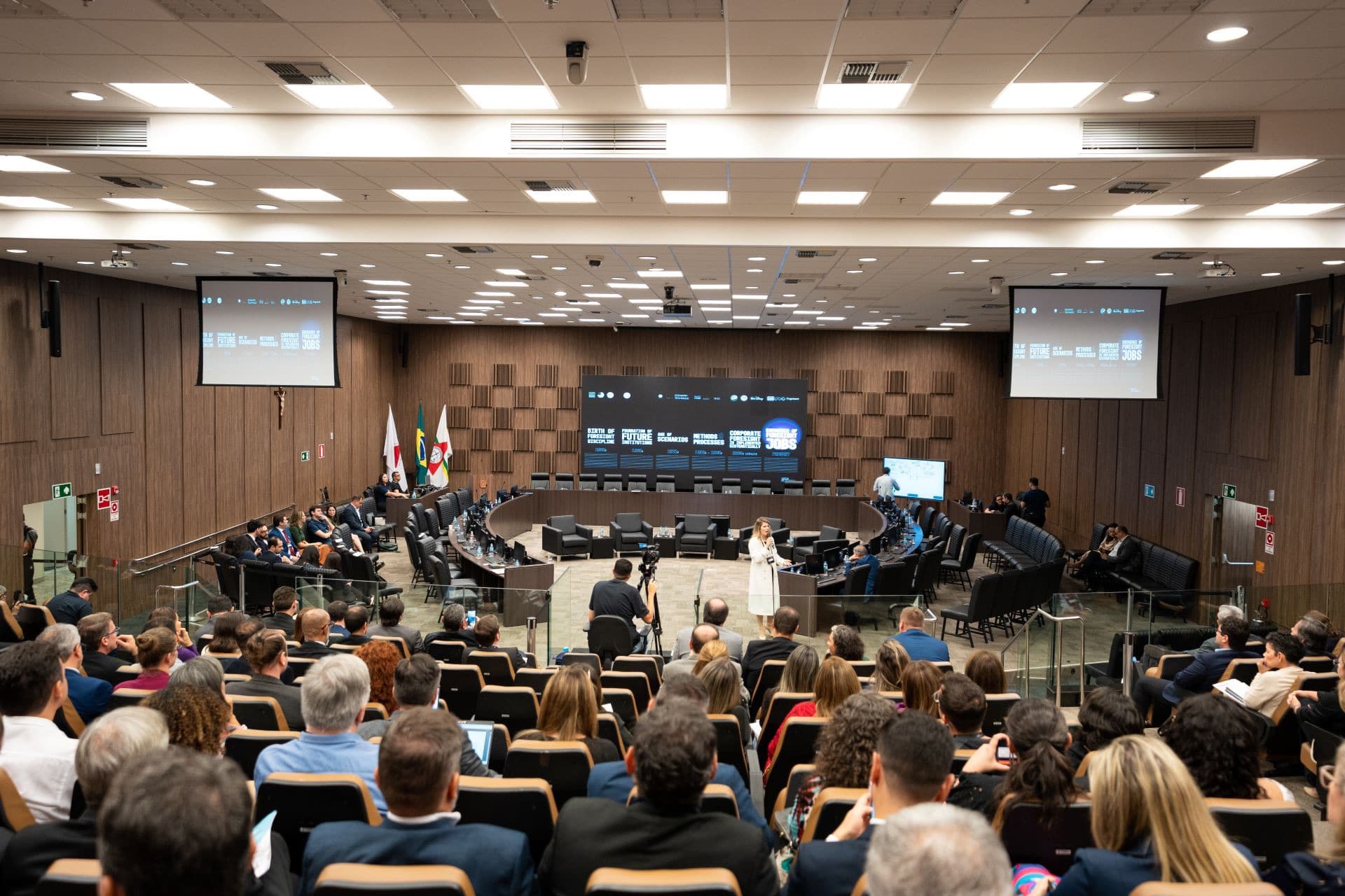 Plenária com participantes assistindo às palestras, com telões ao fundo