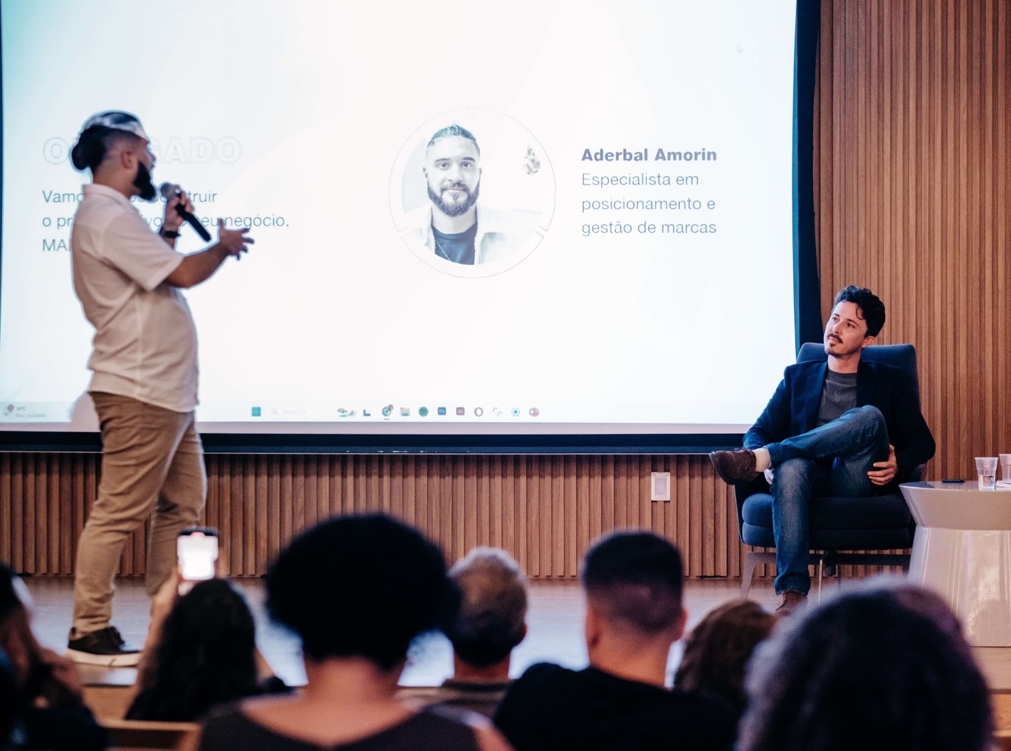 Mediador de pé conduz o talk-show enquanto o convidado responde sentado, com a apresentação no telão