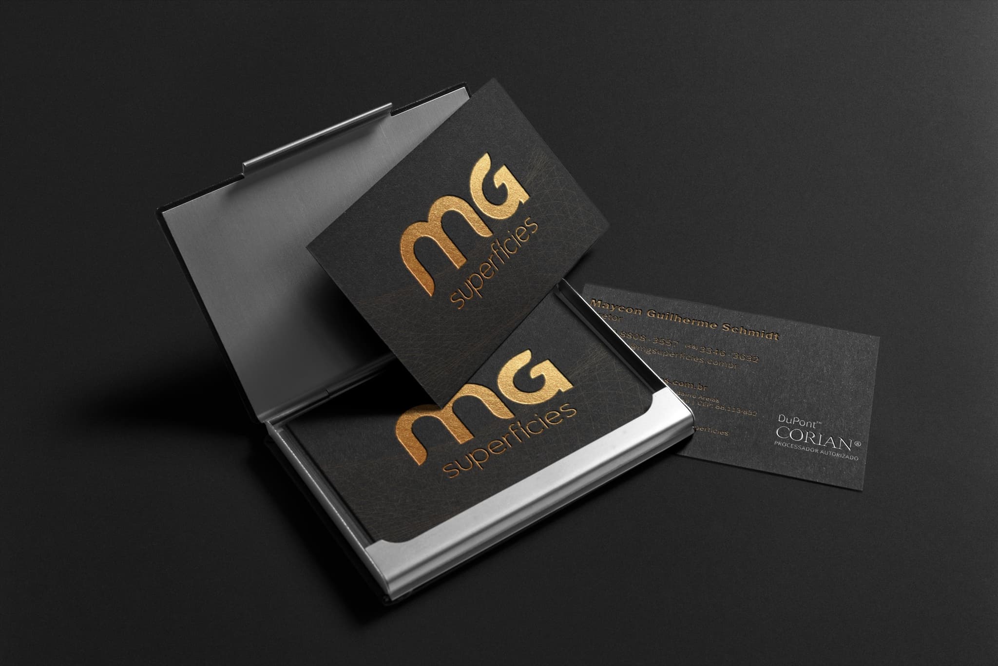 Cartões de visita da MG Superfícies em porta-cartões metálico sobre fundo preto: o monograma dourado em hot stamping sobre papel texturizado, com o verso trazendo o selo de processador autorizado DuPont Corian