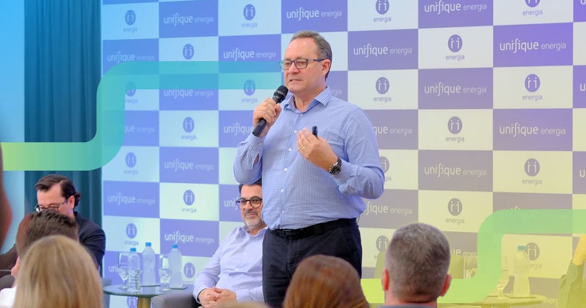 Ruy, da UNIFIQUE, fala ao microfone para o público diante de um painel com a marca unifique energia