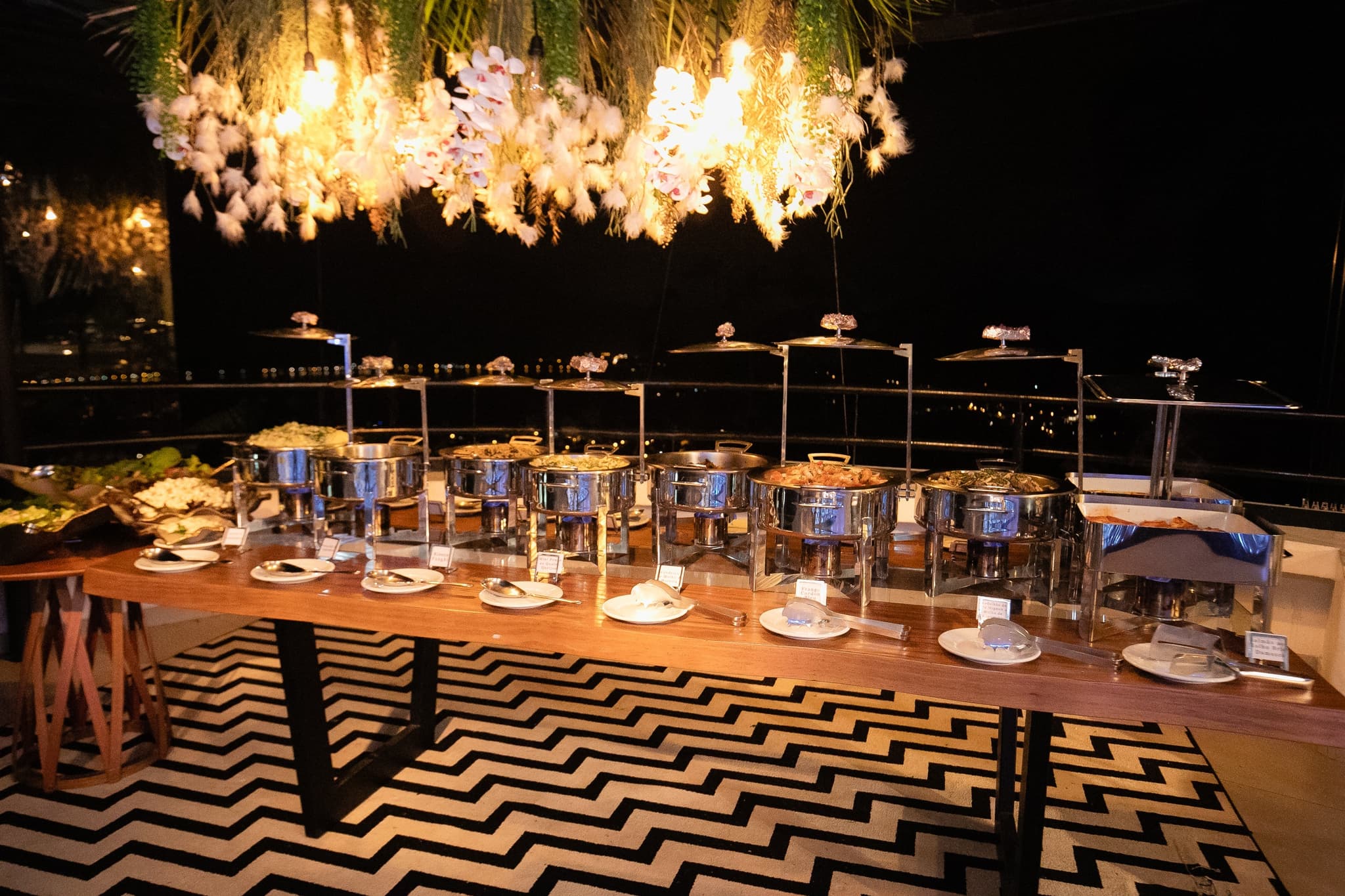 Mesa de buffet com réchauds sob arranjo de flores suspenso e piso de padrão geométrico